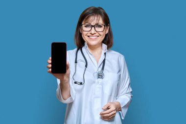 Woman doctor in white uniform with stethoscope showing smartphone screen to camera, on blue color background. Medicine technology applications health care, mockup banner telemedicine