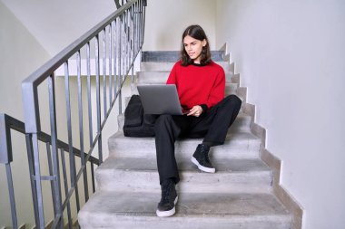 Sırt çantalı yakışıklı bir öğrencinin portresi. Binanın içinde dizüstü bilgisayar kullanıyor, görüntülü konuşuyor. Gençlik, kolej, üniversite, eğitim, teknoloji, yaşam tarzı, gençlik kavramı