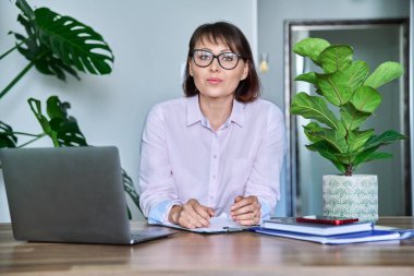 Orta yaşlı, gülümseyen bir iş kadınının portresi masada dizüstü bilgisayarla oturuyor. Gözlüklü güzel olgun bir kadın kameraya bakıyor, iş yerinde çalışıyor, serbest çalışıyor, 40 'lı yaşlarda insanlar konsepti var.