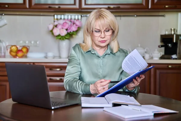 Senior business woman executive working remotely using laptop for video call conference chat sitting at home at table in kitchen. Online meeting with colleagues clients, technology business 50s people