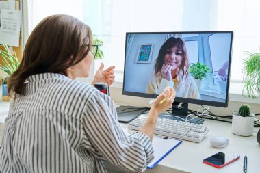 Mature woman talking online with teenage girl using video call on computer, home interior. Virtual meeting, therapy session with psychologist. Chat conference technology psychology education learning