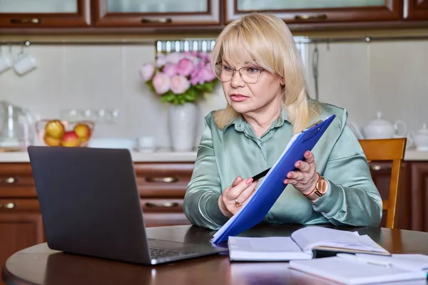 Senior business woman executive working remotely using laptop for video call conference chat sitting at home at table in kitchen. Online meeting with colleagues clients, technology business 50s people