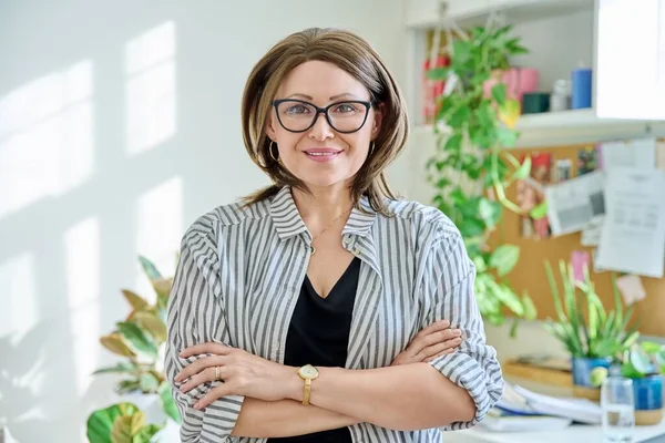 Portrait of confident mature woman looking at camera at home. Middle aged smiling successful female in glasses with crossed arms. 40s age, lifestyle, people concept