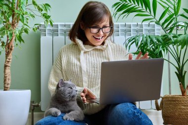 Middle aged woman looking in laptop webcam, making video conference call talking online at home. Mature female in warm clothes sitting with cat near heating radiator in winter autumn season, blog vlog