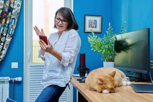 Middle aged woman in home office using smart phone for video call chat conference, pet ginger cat sleeping on desk. Home, animals, technology, lifestyle, work, people concept