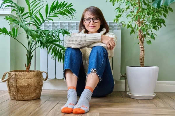 Winter home portrait of middle aged woman in warm knitwear sweater looking at camera. Mature female warming near heating radiator. Cold autumn winter season, leisure lifestyle, 40s people concept