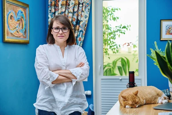 Confident middle aged woman in home office looking at camera. Portrait of mature smiling female with crossed arms, near table with computer pet cat sleeping on desk. Freelance, remote work at home