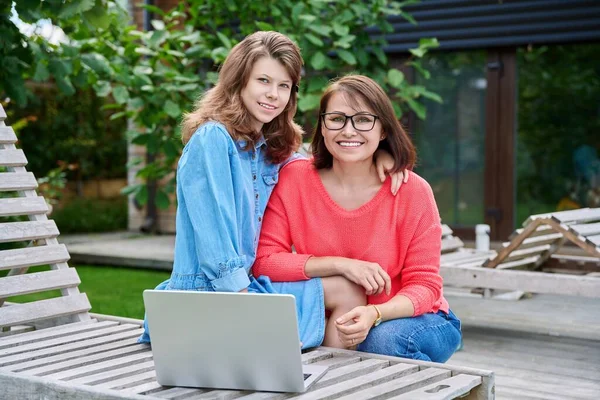 Portrait of happy mom and teenage daughter looking at camera. Smiling hugging mother and child together in backyard on wooden outdoor chairs with laptop. Family, parent child, mothers day, lifestyle