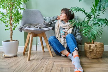 Cold winter season woman with cat near heating radiator. Middle-aged female in warm woolen knitted sweater with cup of hot drink and pet lying on chair. Heating season, lifestyle, crisis energy saving