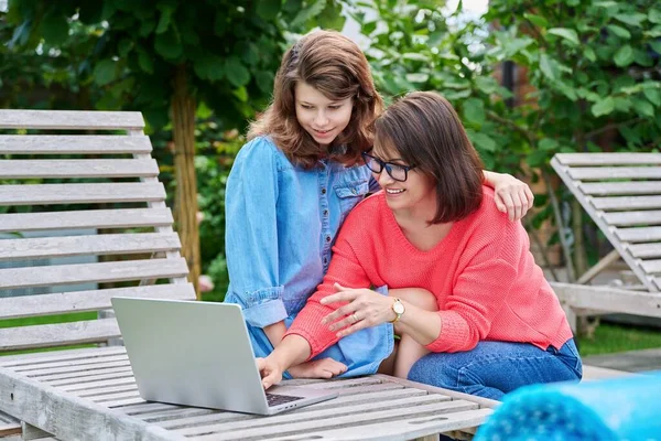 Middle aged mom and preteen daughter looking at laptop together, outdoor on armchairs in backyard. Parent and child hugging using laptop to study leisure shopping