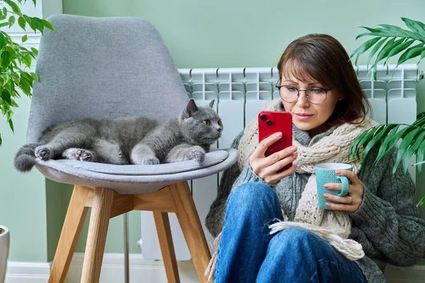 Middle aged woman resting at home in warm clothes using smartphone, warming herself with pet cat near home heating radiator. Winter autumn heating season, lifestyle leisure save energy, crisis concept