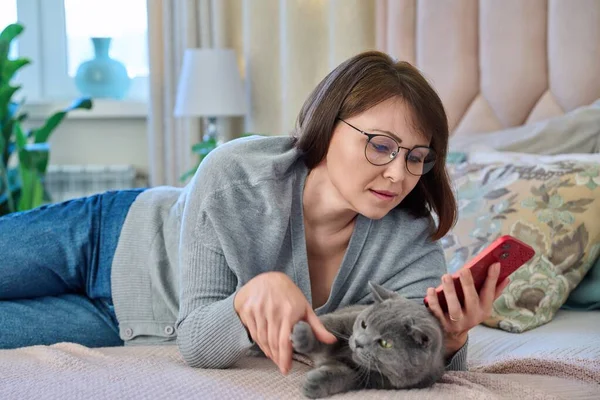 中年齢のリラックスした女性が自宅のベッドに横になり 猫はレジャー読書ビデオのためのスマートフォンのモバイルアプリを使用しています ライフスタイル テクノロジー ペット 人々の概念 — ストック写真