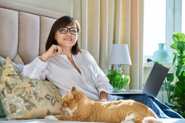 Portrait of business middle aged woman working from home using laptop looking at camera sitting on bed. Beautiful 40s female in glasses with sleeping cat pet cozy home interior remote work freelancing