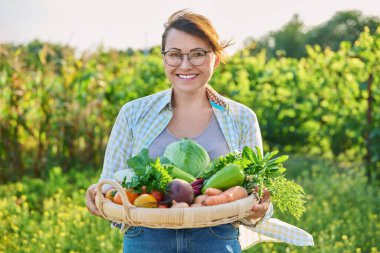 Portrait of smiling middle aged woman with basket of different fresh raw vegetables and herbs, summer nature vegetable garden background. Harvest from organic farm, healthy food nutrition, gardening