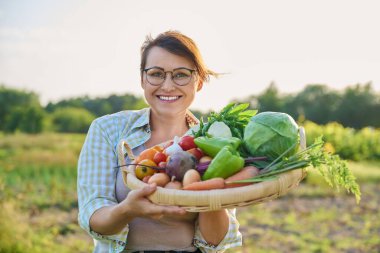 Portrait of smiling middle aged woman with basket of different fresh raw vegetables and herbs, summer nature vegetable garden background. Harvest from organic farm, healthy food nutrition, gardening