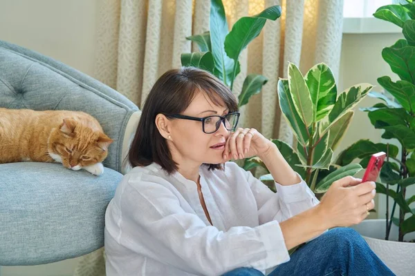 Middle aged woman sitting on floor using smartphone. Female in home interior with sleeping pet ginger red cat on armchair resting with phone. Lifestyle leisure, animal, mature people, comfort calmness