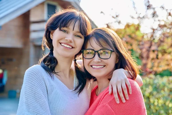 Portrait of happy mom and teenage daughter looking at camera. Smiling hugging mother and teen girl together outdoor in backyard. Family, parent and teenager, mothers day, lifestyle concept