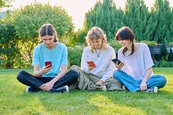 一群青少年坐在草地上 拿着智能手机 三位青少年朋友在草坪上放松 夏日阳光明媚 用手机应用程序休闲 生活方式 技术和友谊 — 图库照片