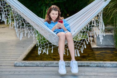 Hamakta oturan küçük kız boş zamanlarında iletişim için akıllı telefon kullanıyor. 12-13 yaşlarında arka bahçedeki hamakta dinleniyor.