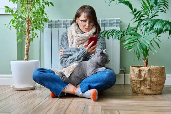 Middle aged woman resting at home in warm clothes using smartphone, warming herself with pet cat near home heating radiator. Winter autumn heating season, lifestyle leisure save energy, crisis concept