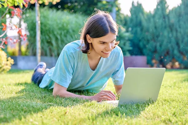 Teenage male lying on grass using laptop. Guy teenager student 18, 19 years old on lawn, technologies for studying leisure communication shopping in backyard in garden. Lifestyle, youth concept