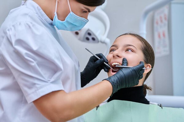 Young teenage female at dental checkup in clinic. Teenage girl sitting in chair, doctor dentist with tools examining patients teeth. Adolescence, hygiene, treatment, dental health care