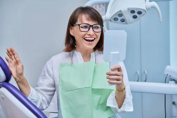 Middle-aged female patient in dental office looking in mirror at treated prosthetic teeth. Happy smiling woman after prosthetic treatment, implantation, dental health, beauty care concept