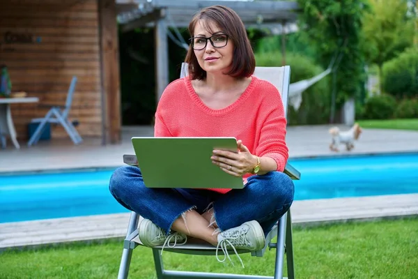 Middle aged woman sitting in chair in backyard with laptop. Female using laptop for work and leisure, pool house lawn background. Lifestyle technology work relax 40s people