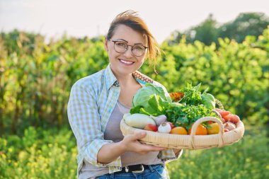 Portrait of smiling middle aged woman with basket of different fresh raw vegetables and herbs, summer nature vegetable garden background. Harvest from organic farm, healthy food nutrition, gardening
