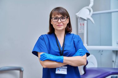 Ofisteki kadın doktor portresi. Kendine güveni tam orta yaşlı bir kadın kameraya bakıyor ve dişçi koltuğunun yanında çapraz kolları var. Diş hekimliği, tıp, uzman, kariyer, diş sağlığı konsepti