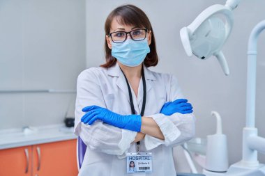 Ofiste yüz maskesi takmış ciddi bir kadın dişçinin portresi. Kendine güveni tam orta yaşlı bir kadın kameraya bakıyor ve dişçi koltuğunun yanında çapraz kolları var. Diş hekimi, uzman, kariyer, sağlık hizmetleri