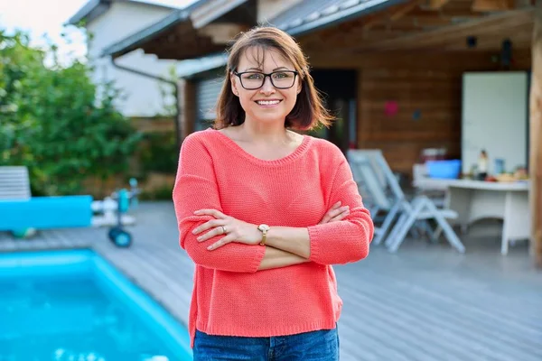 Portrait of smiling middle aged woman looking at camera outdoor. Successful confident mature woman with crossed arms in backyard of house with swimming pool