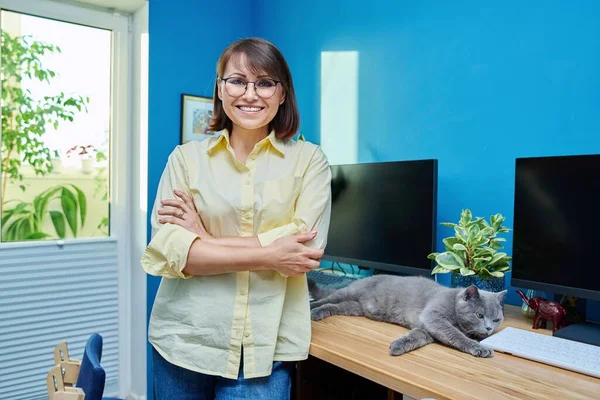 Confident middle aged woman in home office looking at camera. Portrait of mature smiling female with crossed arms, near table with computer pet cat sleeping on desk. Freelance, remote work at home