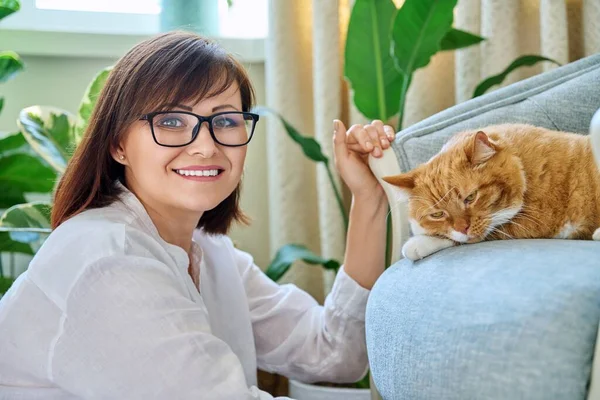 Portrait of smiling 40s woman with pet ginger cat lying on armchair in home interior. Beautiful happy middle aged female looking at camera. Lifestyle, animal, mature people, comfort happiness calmness
