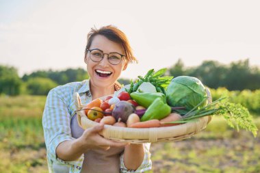 Portrait of smiling middle aged woman with basket of different fresh raw vegetables and herbs, summer nature vegetable garden background. Harvest from organic farm, healthy food nutrition, gardening