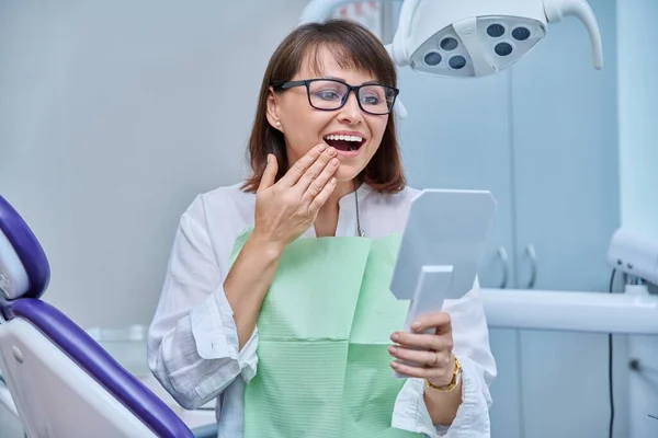Middle-aged female patient in dental office looking in mirror at treated prosthetic teeth. Happy smiling woman after prosthetic treatment, implantation, dental health, beauty care concept