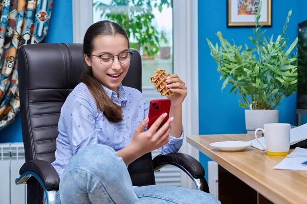Adolescente Sentada Uma Poltrona Perto Computador Olhando Smartphone Comendo Waffle — Fotografia de Stock