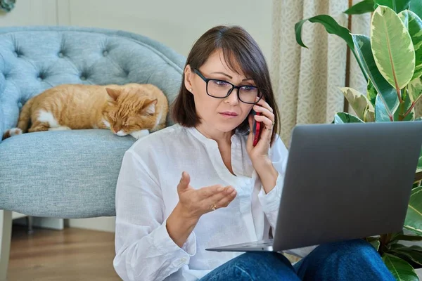 Middle aged woman sitting at home with laptop, working remotely with pet red ginger cat sleeping on armchair. Female freelancer using smartphone laptop, home interior. Technology work people animal