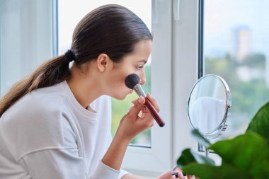 Aynalı genç bir kadın gündüz makyajı yapıyor. Pencere kenarında. Doğal dekoratif kozmetik, genç cilt bakımı, güzellik gençliği konsepti