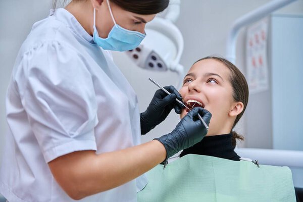 Young teenage female at dental checkup in clinic. Teenage girl sitting in chair, doctor dentist with tools examining patients teeth. Adolescence, hygiene, treatment, dental health care