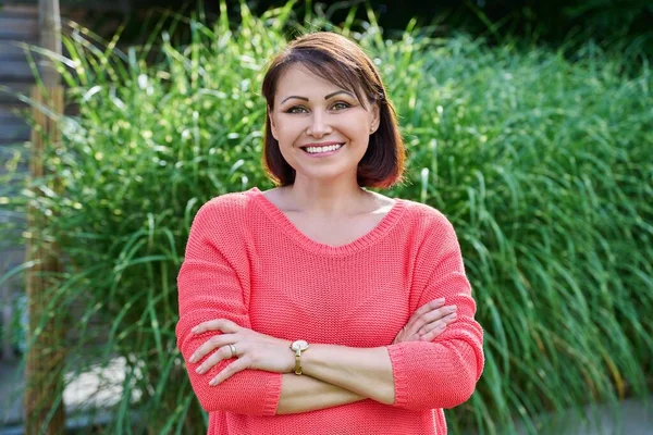 Portrait of smiling middle aged woman looking at camera outdoor. Successful confident mature woman in red with arms crossed, nature green lawn background