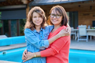 Kameraya bakan mutlu anne ve genç kızın portresi. Arka bahçede anne ve kızı kucaklayan bir gülümseme. Açık havuz kenarında. Aile, ebeveyn ve çocuk, anneler günü, yaşam tarzı konsepti