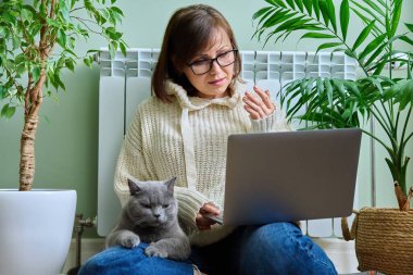 Middle aged woman looking in laptop webcam, making video conference call talking online at home. Mature female in warm clothes sitting with cat near heating radiator in winter autumn season, blog vlog