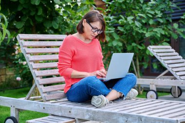 Ahşap iskemlede oturan olgun bir kadın boş zaman ve iş için dizüstü bilgisayar kullanıyor, bir kadın arka bahçede dizüstü bilgisayara bakıyor. Serbest çalışma, uzaktan çalışma, rahatlama, orta yaşlı insanlar konsepti