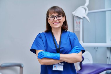 Ofisteki kadın doktor portresi. Kendine güveni tam orta yaşlı bir kadın kameraya bakıyor ve dişçi koltuğunun yanında çapraz kolları var. Diş hekimliği, tıp, uzman, kariyer, diş sağlığı konsepti