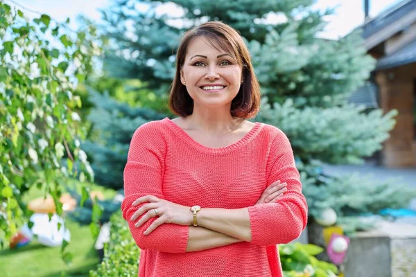 Portrait of smiling middle aged woman looking at camera outdoor. Successful confident mature woman in red with arms crossed, nature green lawn background