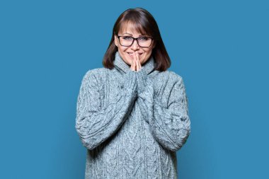 Kendine güvenen orta yaşlı bir kadının mavi renkli stüdyo arkaplanındaki kameraya bakışının portresi. Kış süveteri örmüş olgun bir kadın. Güzellik, kış mevsimi kış mevsimi, 40 'lı yılların insanları konsepti