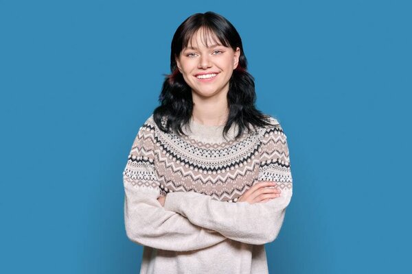 Портрет молодой женщины, смотрящей в камеру в зимнем свитере на фоне студии голубого цвета. Уверенная улыбающаяся женщина 18 лет, 19 лет со скрещенными руками. Молодежь, осень зимний сезон, красота зимой