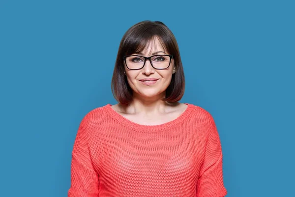 青い色のスタジオの背景にカメラを見て笑顔の中年女性 赤のカジュアルな眼鏡の魅力的な成熟した女性 積極性 ライフスタイル — ストック写真