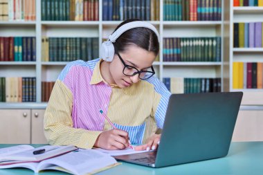 Kütüphanede okuyan genç kız öğrenci. Kulaklıklı bir kadın dizüstü bilgisayar kullanıyor, bilgisayara bakıyor, webinar izliyor, not defterine not alıyor. Ergenlik, teknoloji, eğitim, lise konsepti